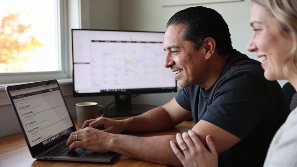 Business owner at a wooden desk in a home office reviewing a list of customer questions on a laptop screen with a content calendar visible in the background
